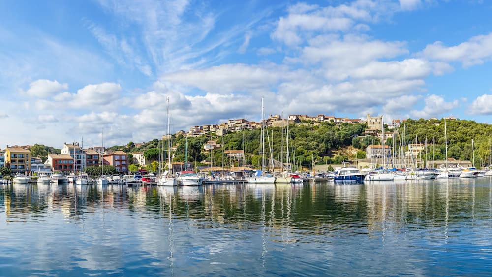 Un port de plaisance avec des bateaux amarrés, une colline parsemée de bâtiments et un ciel dégagé.