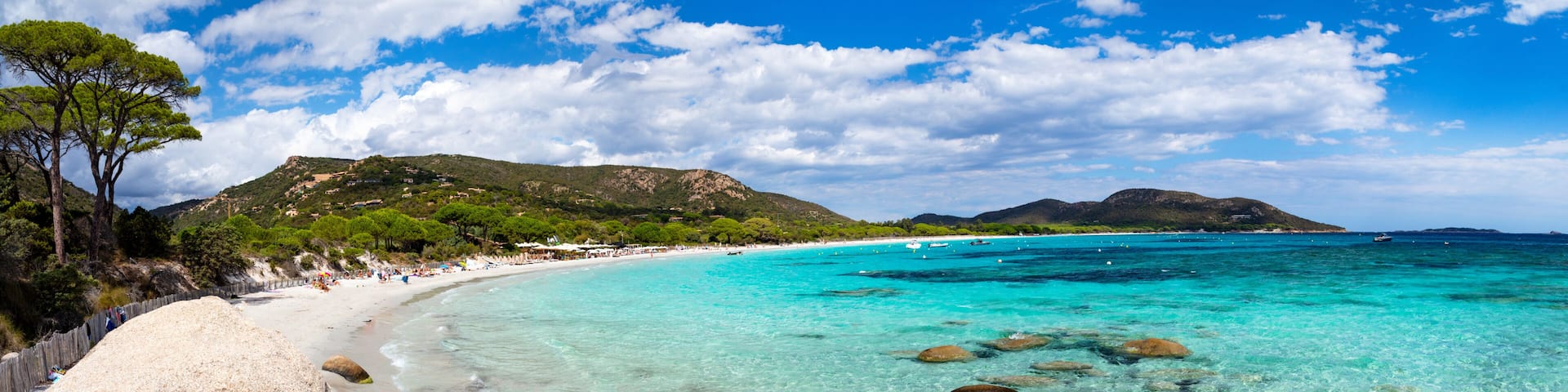 A beach with clear turquoise water, a sandy shore, and a line of rocks leading into the sea.