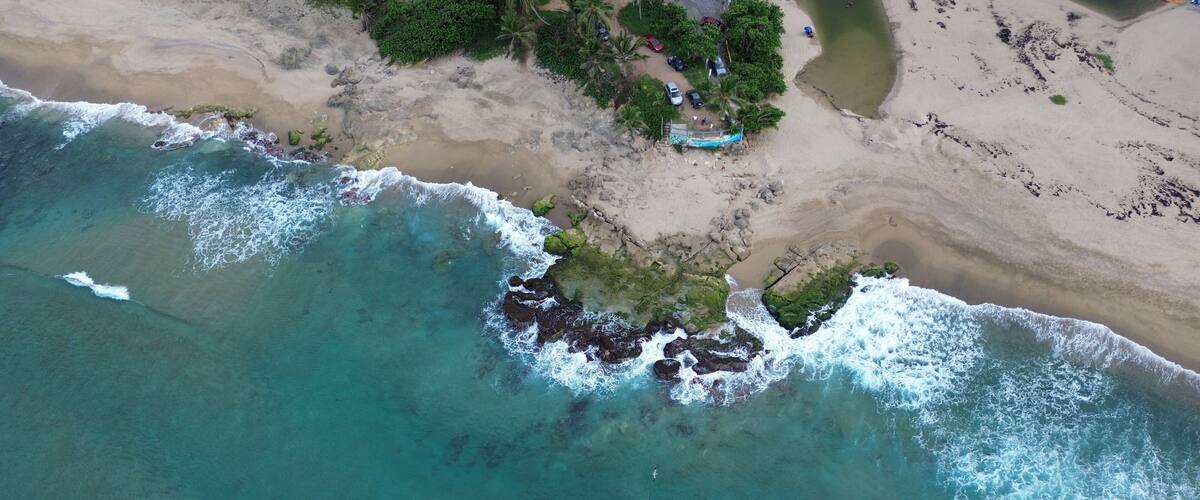 An aerial view of a beach with clear turquoise waters, rocky outcrops, and a small cluster of buildings near the shore.