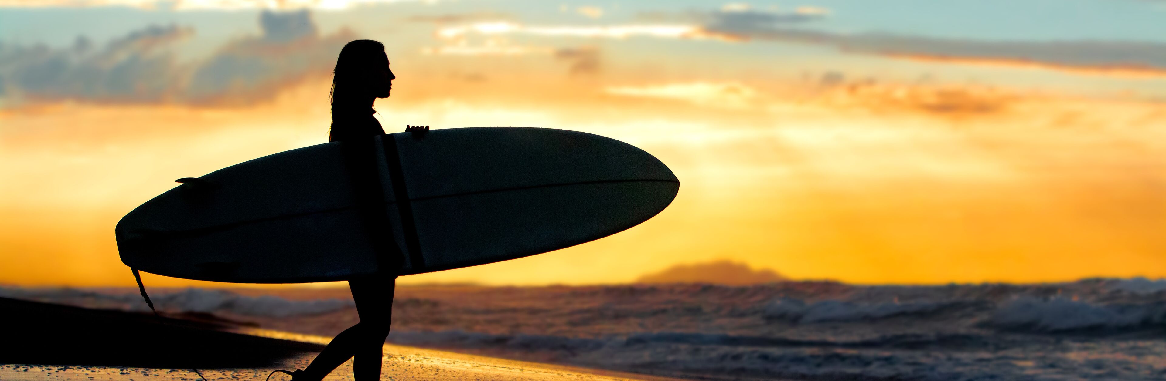 A silhouette of a person holding a surfboard on a beach with waves and a sunset in the background.