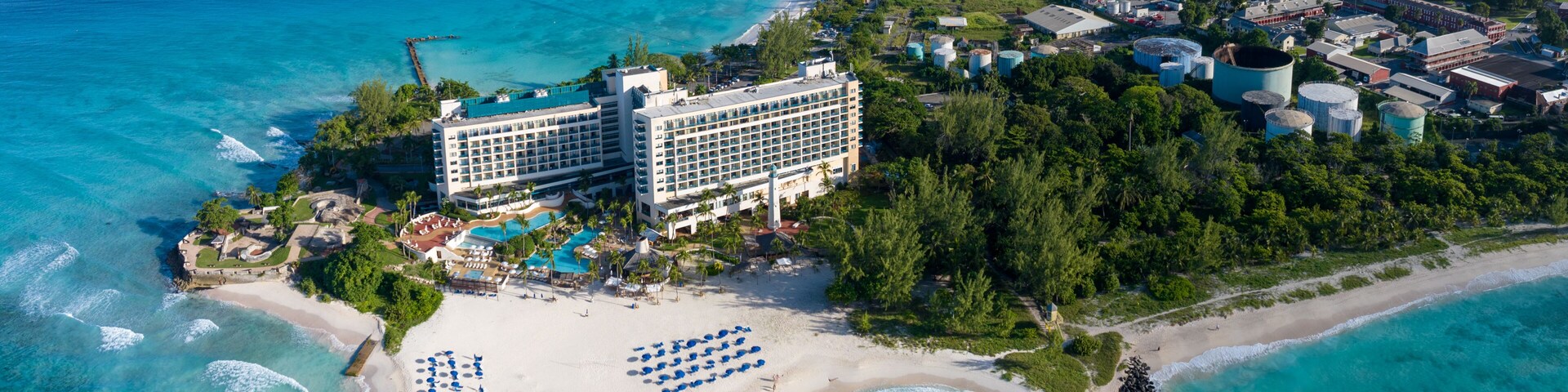 Vista aérea de un complejo turístico costero con un gran hotel, playa y puerto deportivo.