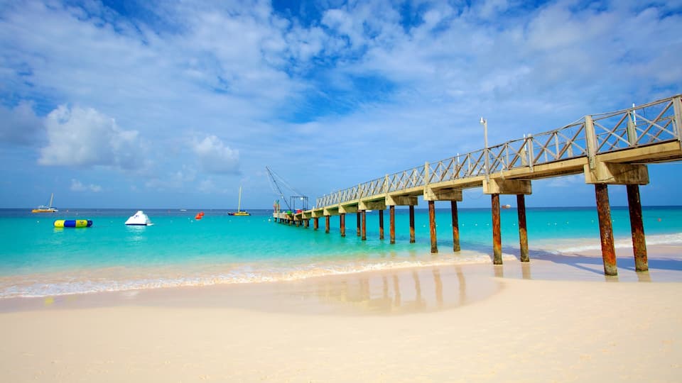 A beach with a wooden pier extending into the turquoise sea, boats, and clear skies.