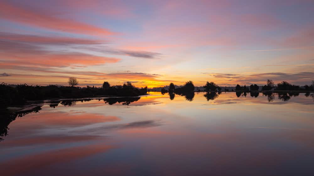 A sunset over a calm lake with silhouettes of trees and a clear reflection.