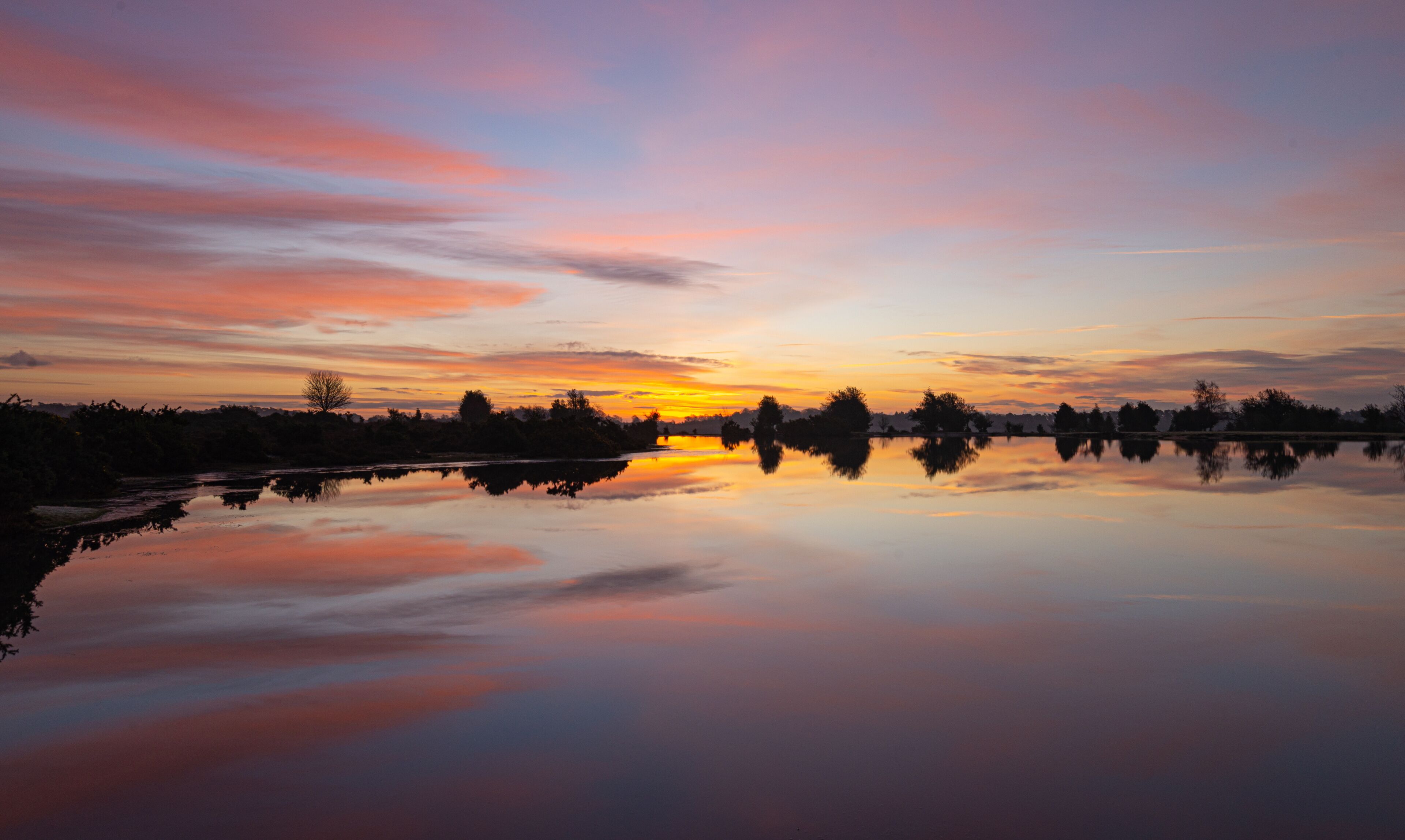 A sunset over a calm lake with silhouettes of trees and a clear reflection.