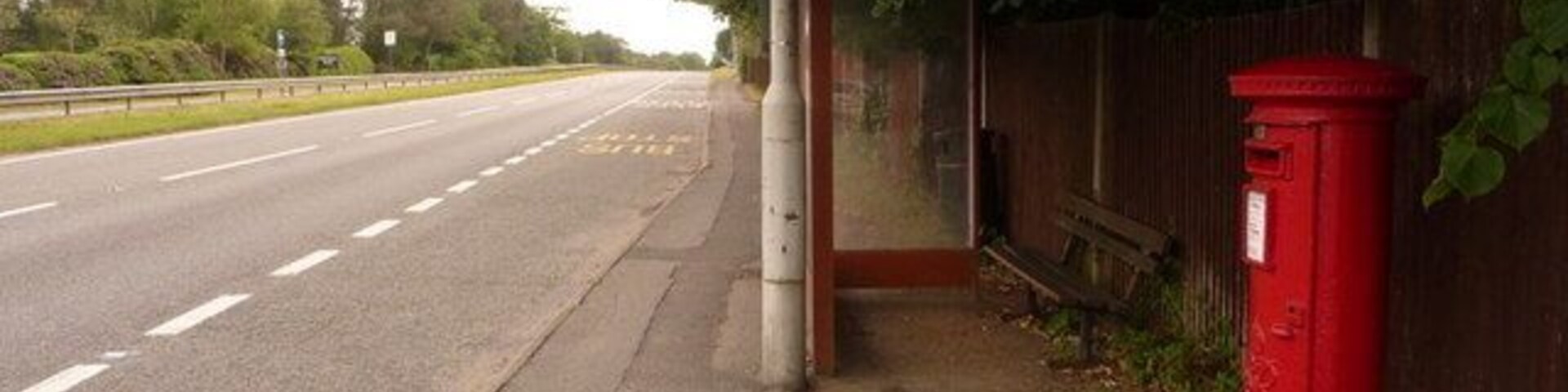 A road with a sidewalk, a red post box, and a bench under a tree.