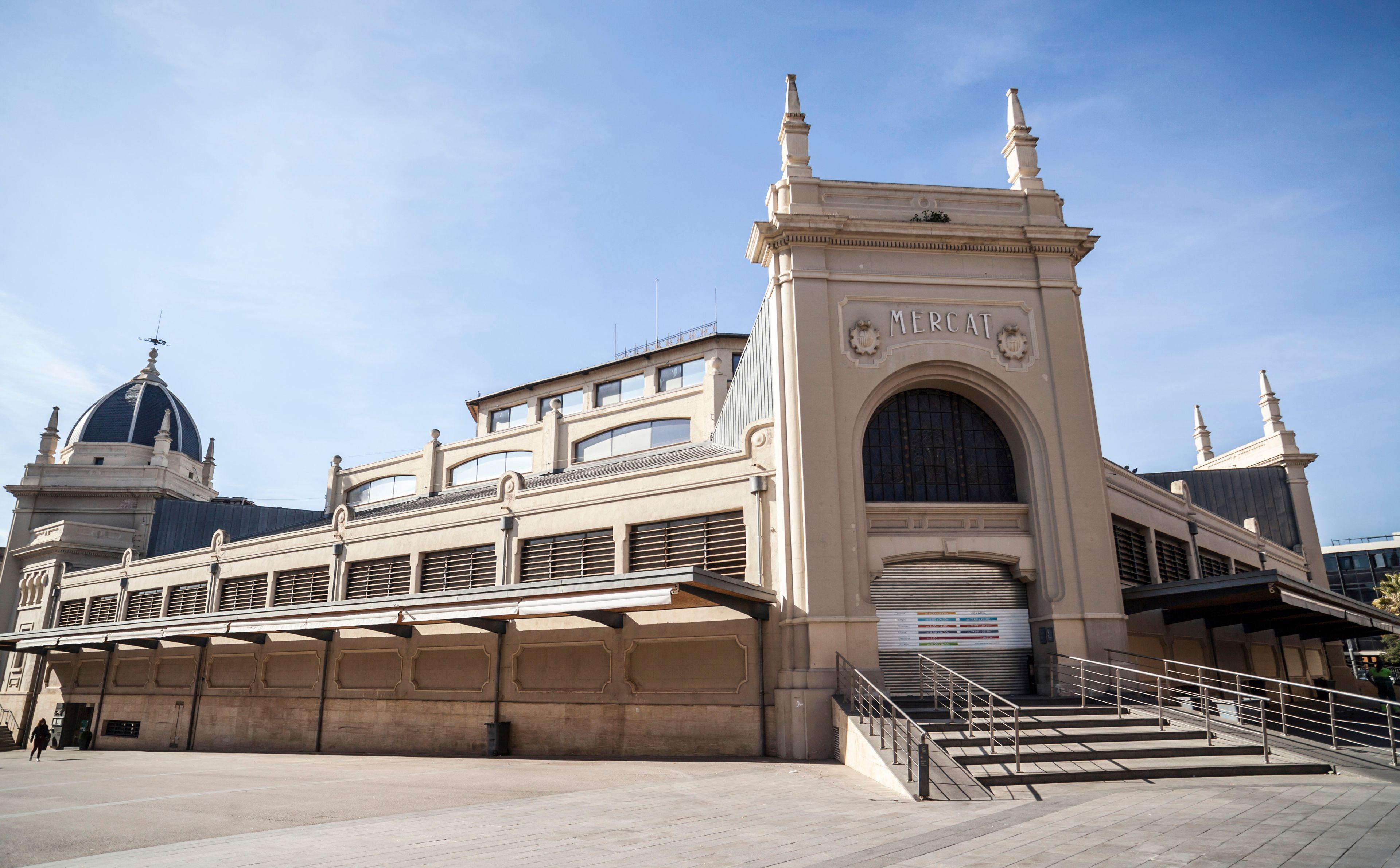 Ein historisches Gebäude mit einem markanten Bogen, auf dem 'MERCAT' steht, und einer Kuppelstruktur im Hintergrund.