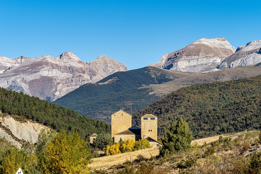 Una iglesia de piedra con dos torres, rodeada por un paisaje montañoso de vegetación escasa y cielos despejados.
