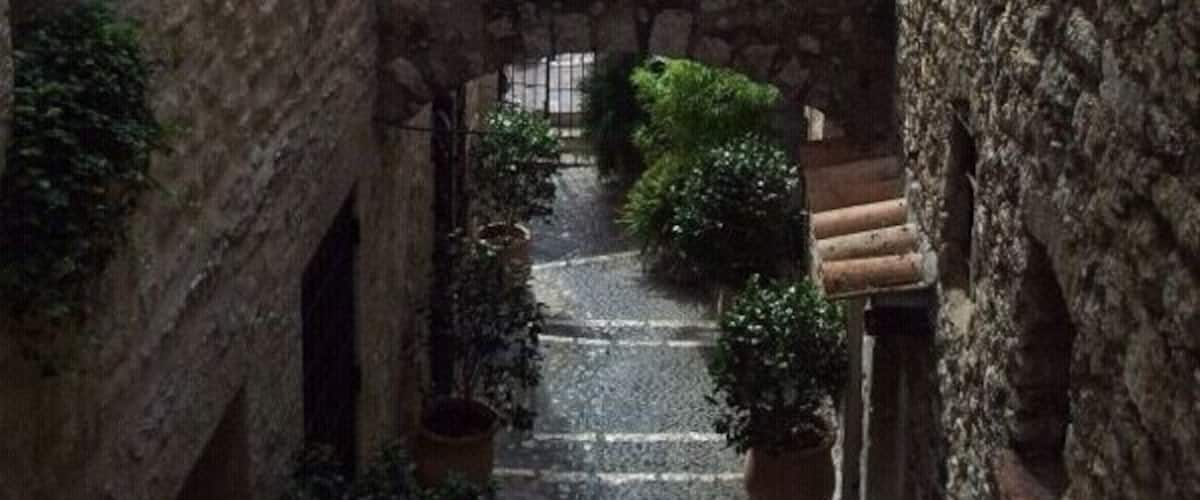 A narrow stone pathway between buildings with a view of a distant town and greenery.