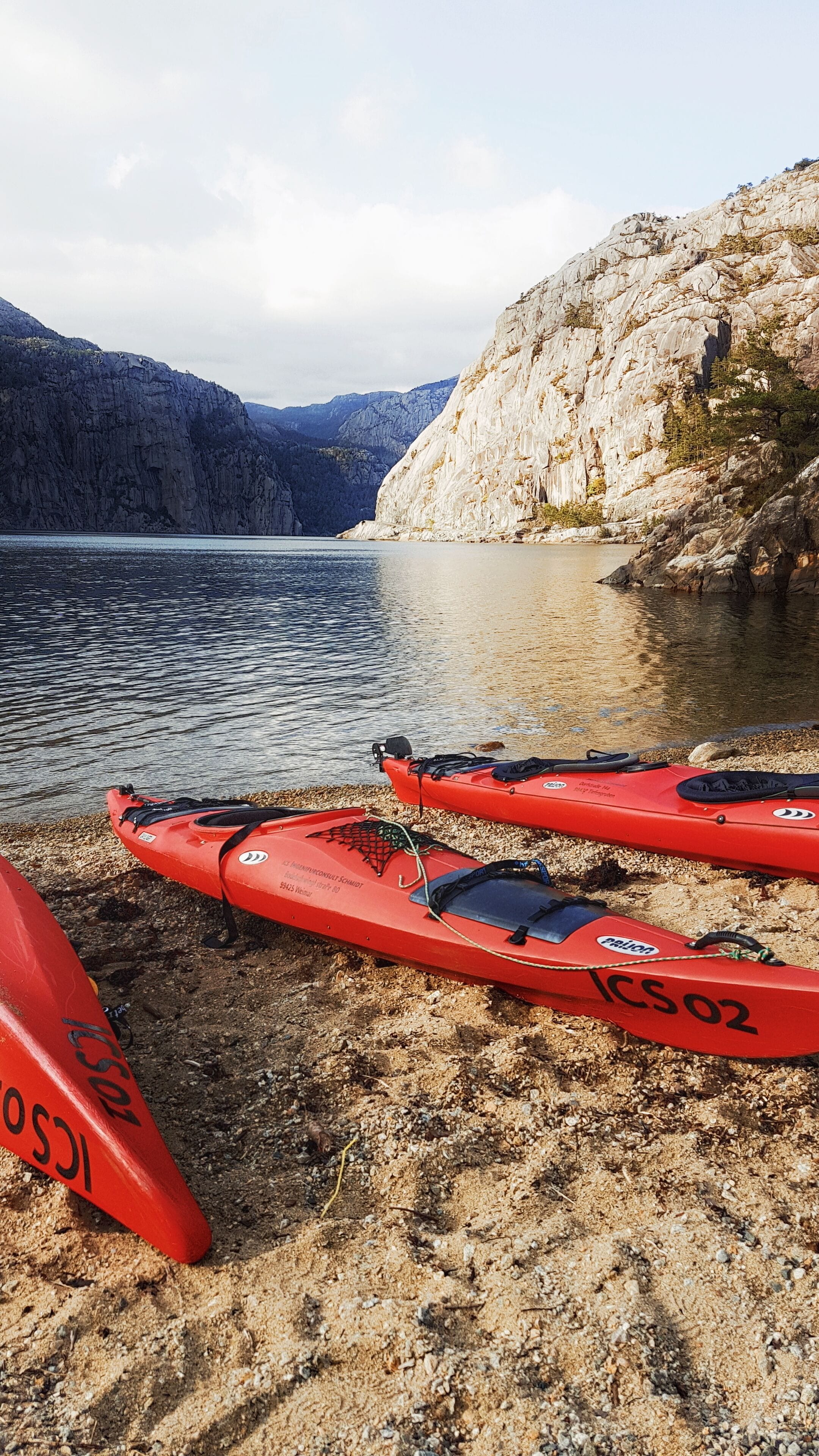 Two red kayaks on a pebble beach with a lake and rocky cliffs in the background.