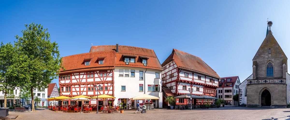 A town square with half-timbered buildings, outdoor cafes, and a church with a tall steeple.