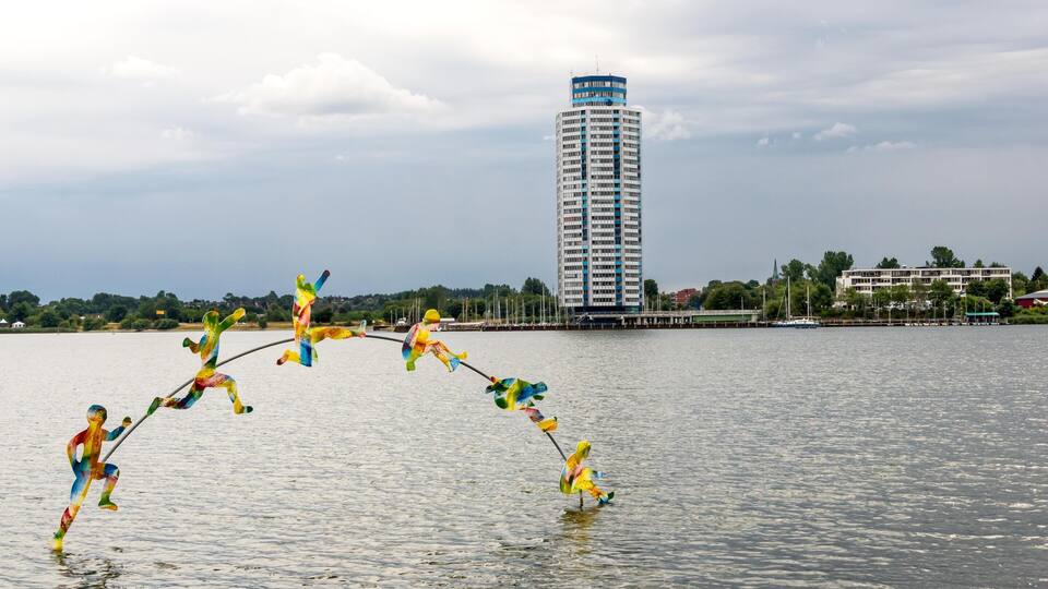 A body of water with colorful kite sculptures, a tall building, and a distant shoreline with trees and buildings.