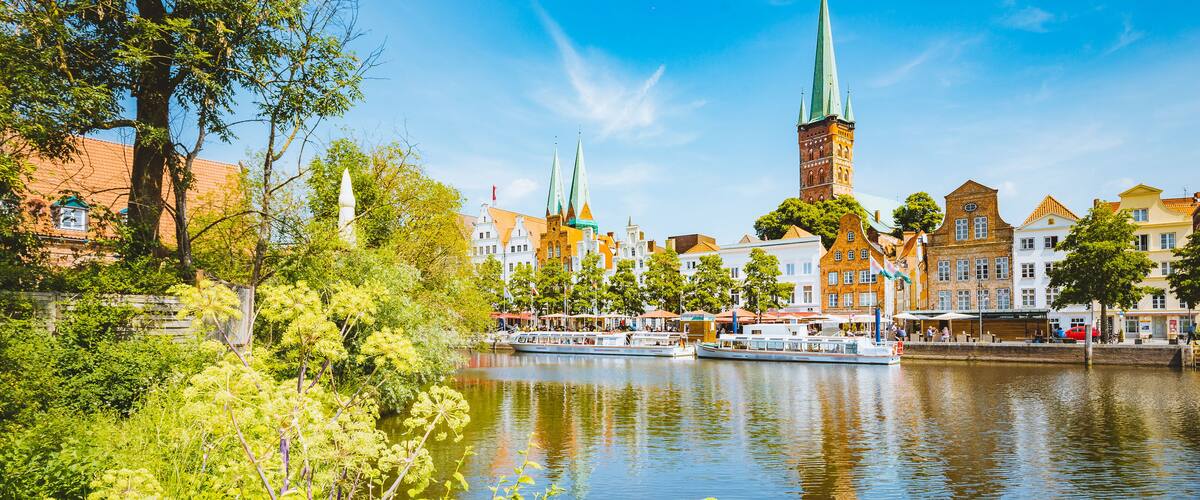 A river with boats, a church with a tall spire, and colorful buildings along the riverbank.