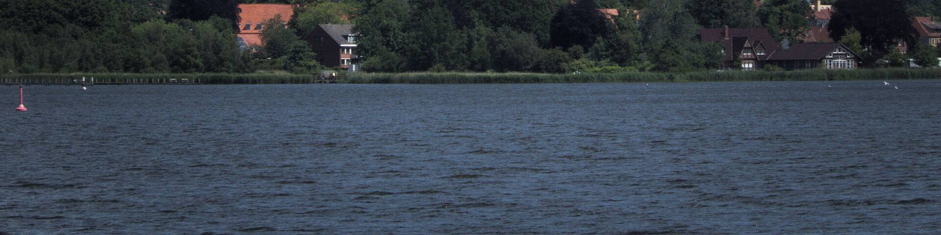 A church with a tall spire and a smaller tower, surrounded by trees and houses across a body of water.