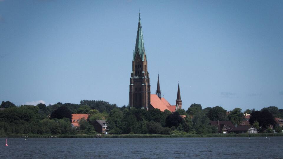 A church with a tall spire and a smaller tower, surrounded by trees and houses across a body of water.