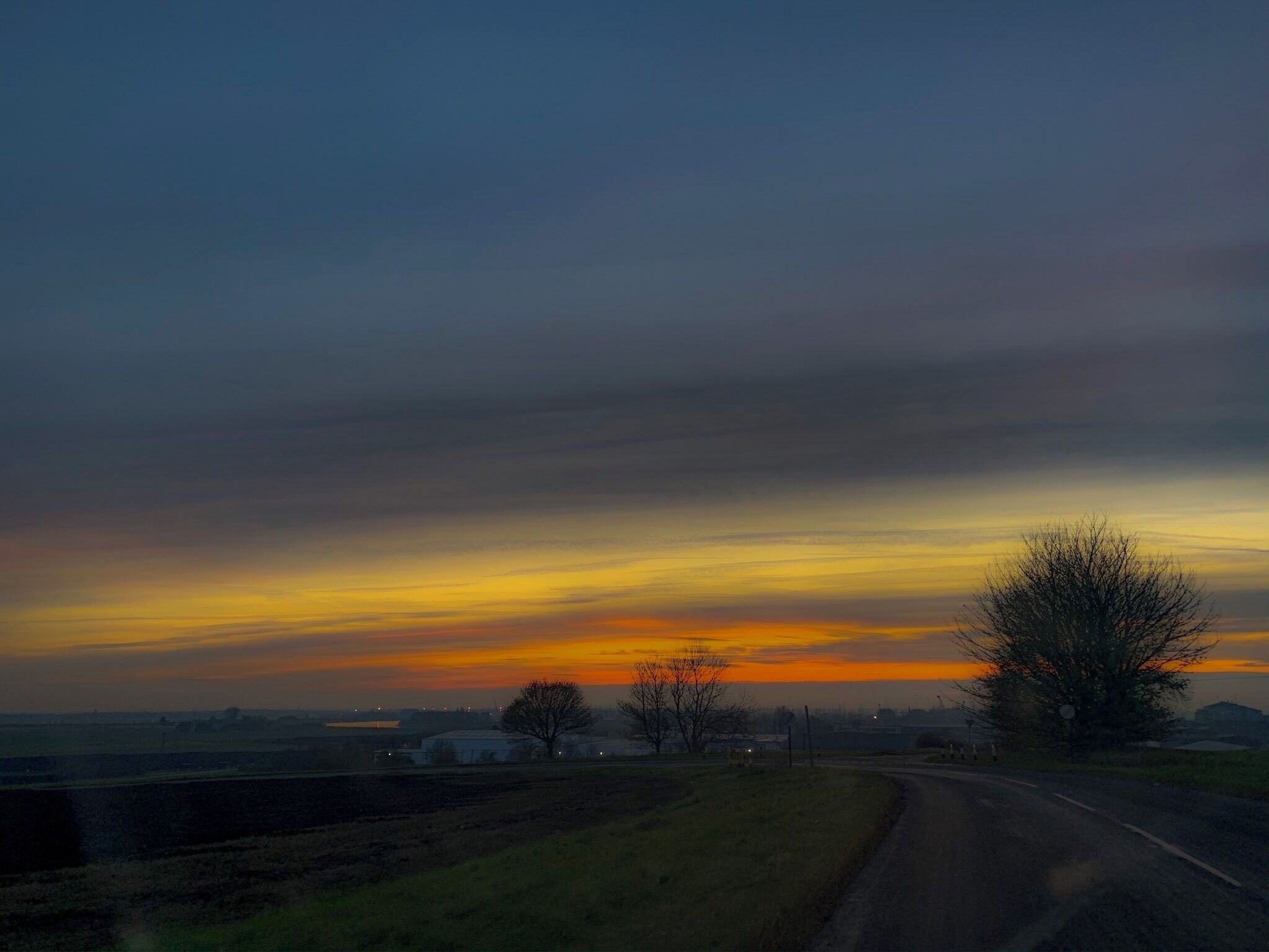 A rural landscape with a road curving to the right, a tree on the right, and a sunset with orange and yellow hues in the sky.