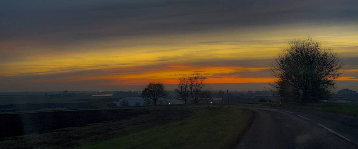 A rural landscape with a road curving to the right, a tree on the right, and a sunset with orange and yellow hues in the sky.