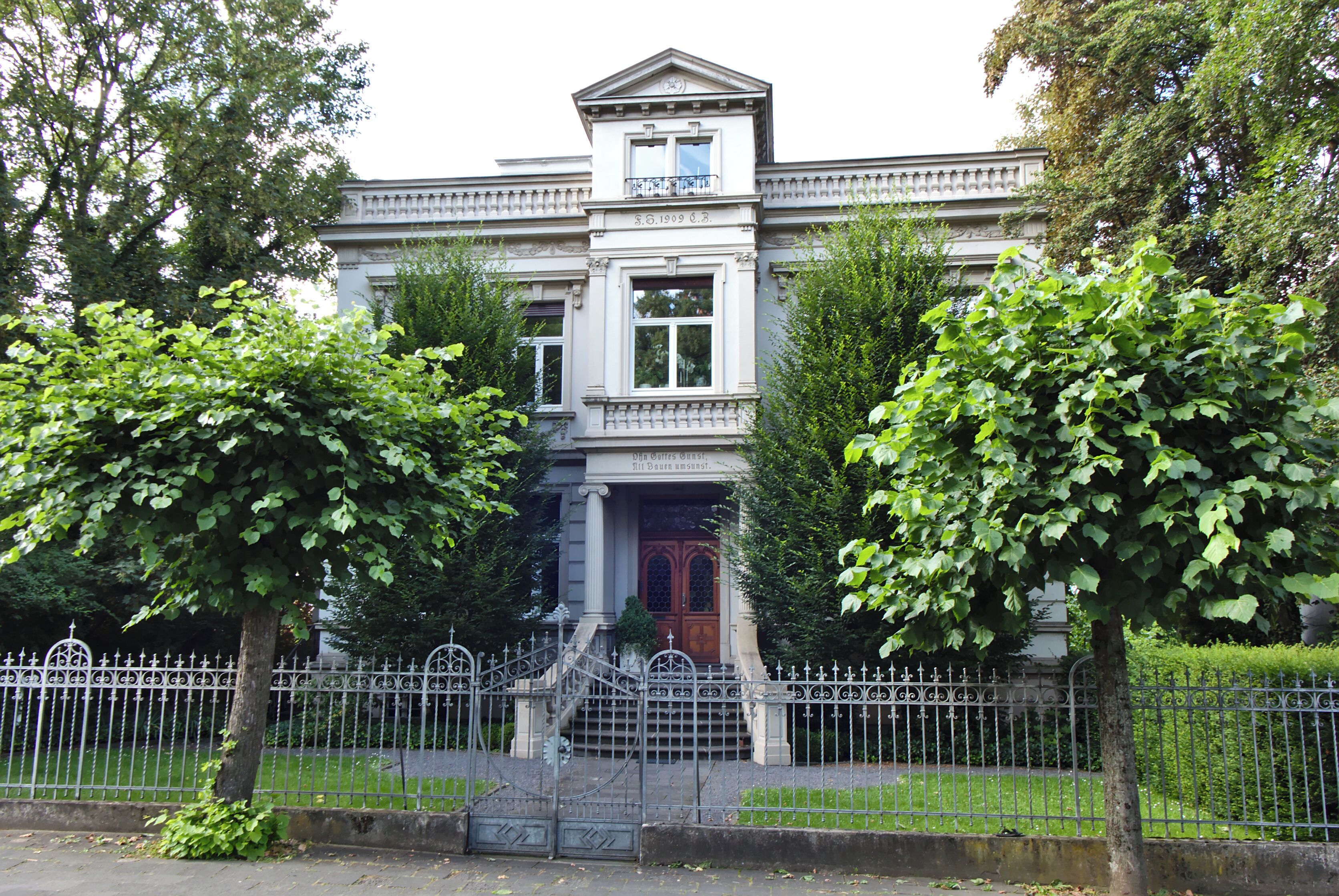 A stately building with a grand entrance, surrounded by lush greenery and a decorative iron fence.