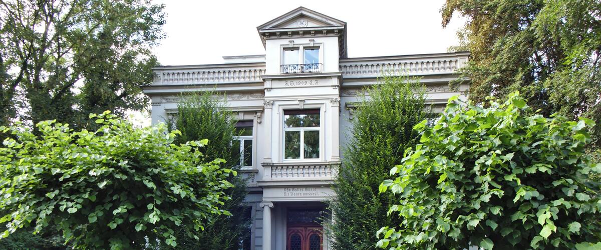 A stately building with a grand entrance, surrounded by lush greenery and a decorative iron fence.