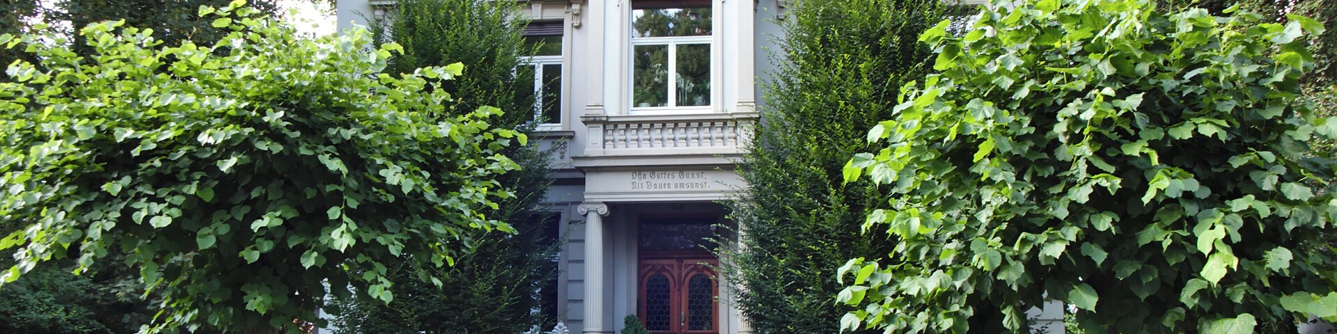 A stately building with a grand entrance, surrounded by lush greenery and a decorative iron fence.