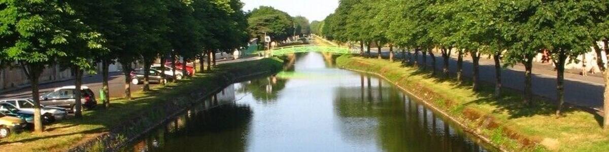 Canal de Berry à Saint-Amand Montrond, Cher, vue O-E. Aménagé entre début et mi-XIXe s. au petit gabarit berrichon (27,5 x 2,6 m, port max. 60 t), il est déclassé en 1955. On est loin des dimensions Freycinet, 38,5 x 5,05 m, port de 250 à 400 t, normes d’une grande part du réseau fluvial français, un peu plus tard ! Ses 3 branches cumulées, représentent 320 km de voies d’eau. Celle traversant la ville, dite sud et qui opère un large coude, vient de Montluçon, Allier.