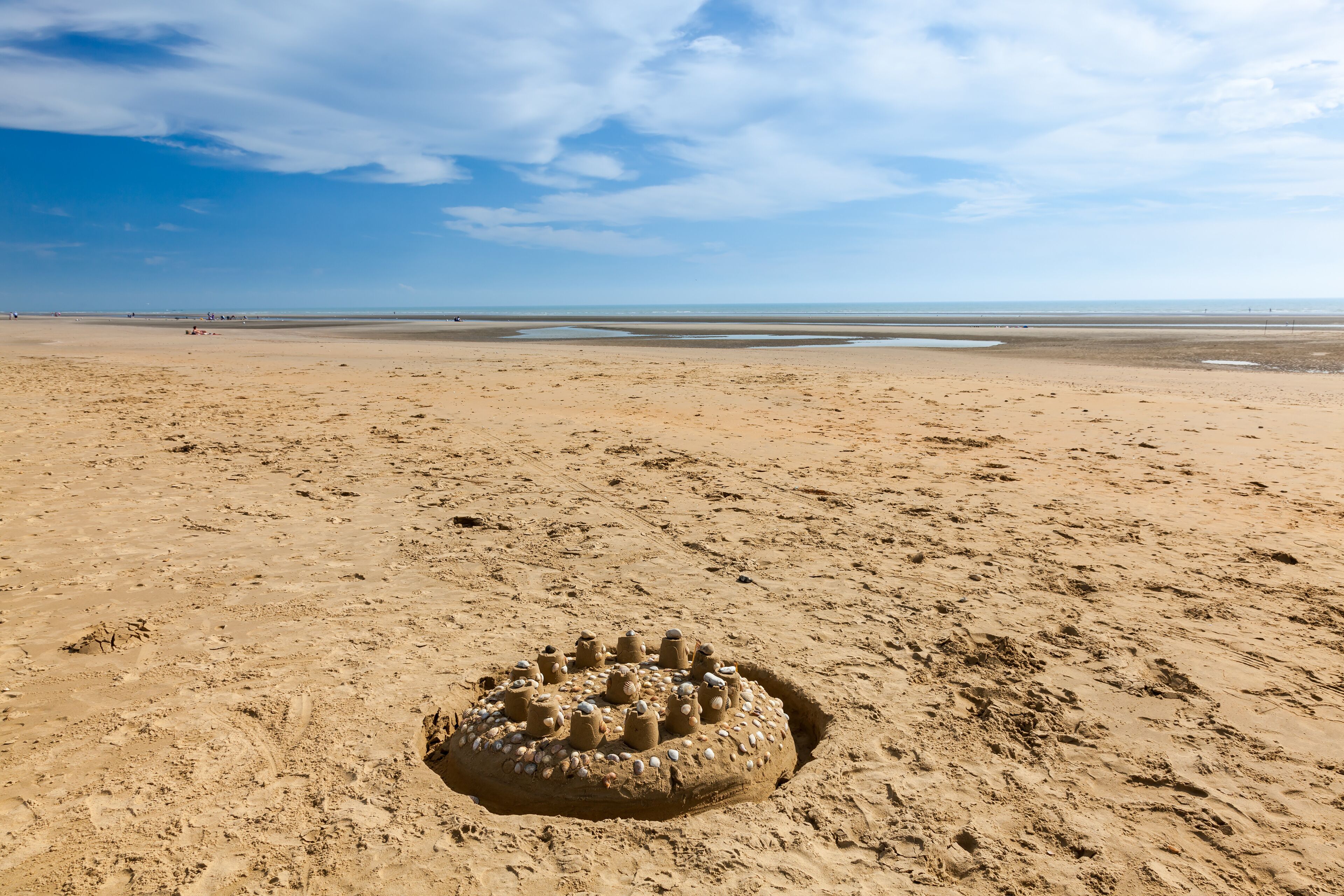 Een zandstrand met een groot zandkasteel op de voorgrond en een rustige zee op de achtergrond.