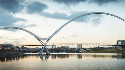 A modern bridge with a unique arch design spanning over a river, with buildings and a clear sky in the background.