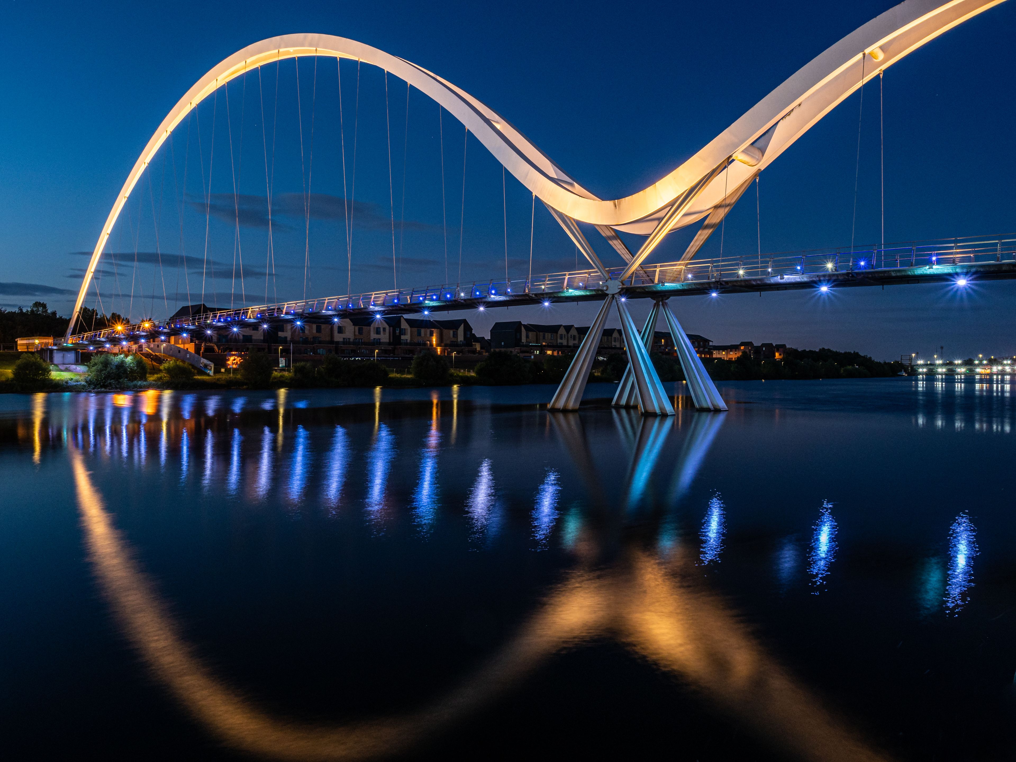 A modern bridge with a distinctive arch design, illuminated lights, and reflections on the water below.
