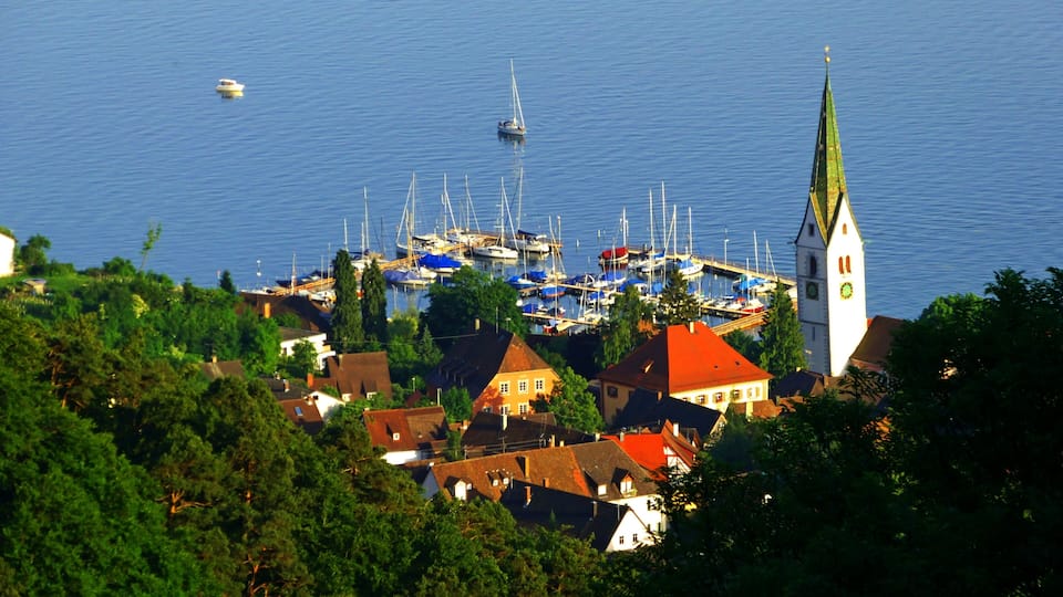 A coastal town with a prominent church tower, a marina with sailboats, and lush greenery.