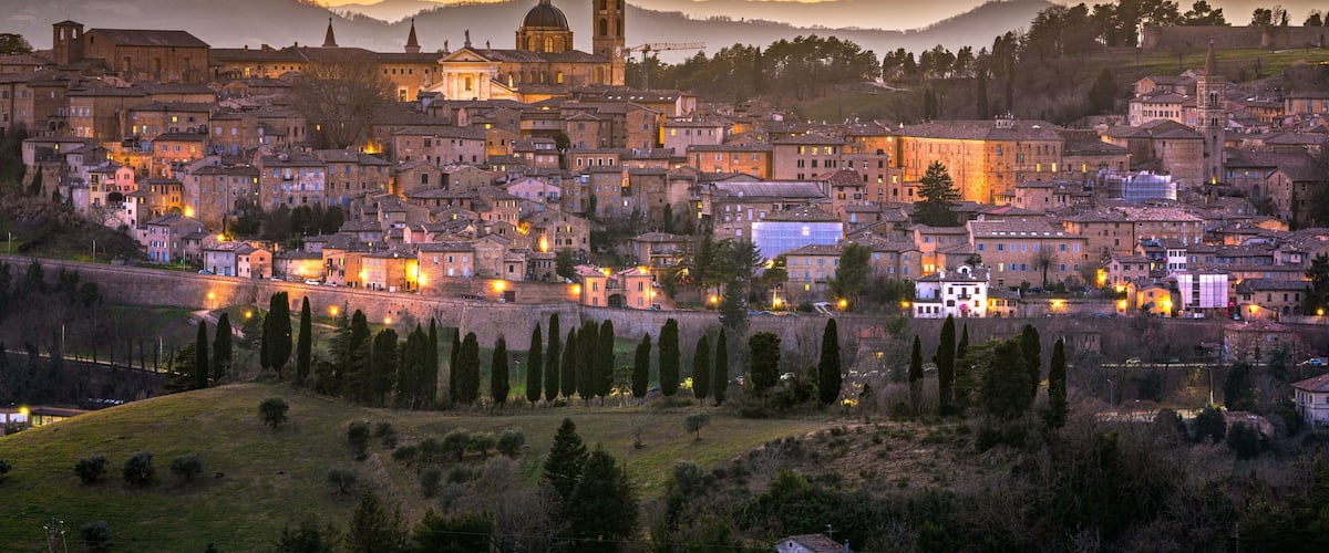 A historic town with a prominent church and bell tower, surrounded by rolling hills and a sunset sky.