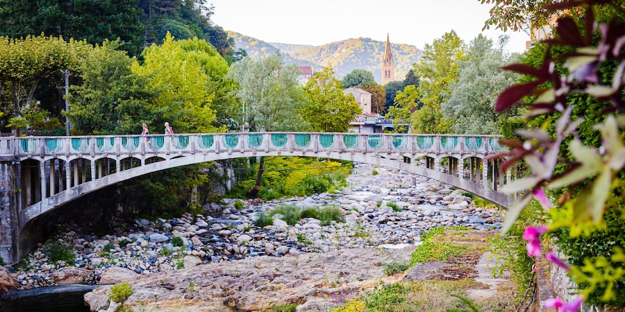 A stone bridge over a rocky river with greenery and a distant church.