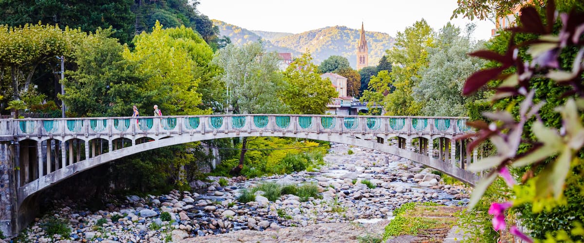 A stone bridge over a rocky river with greenery and a distant church.