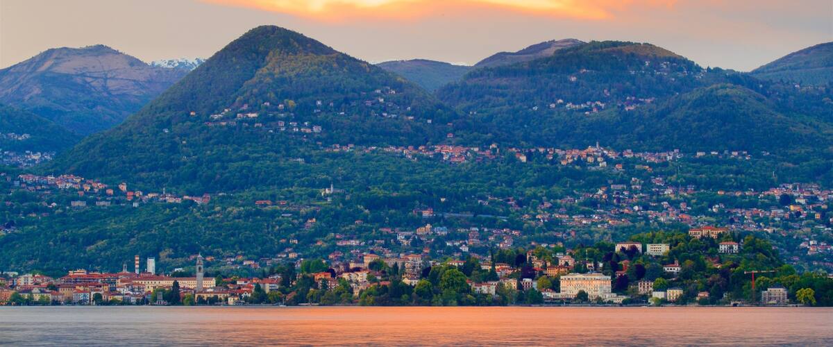 A mountainous landscape with a town at the base, a body of water in the foreground, and a sunset in the background.