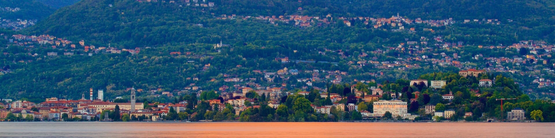 A mountainous landscape with a town at the base, a body of water in the foreground, and a sunset in the background.