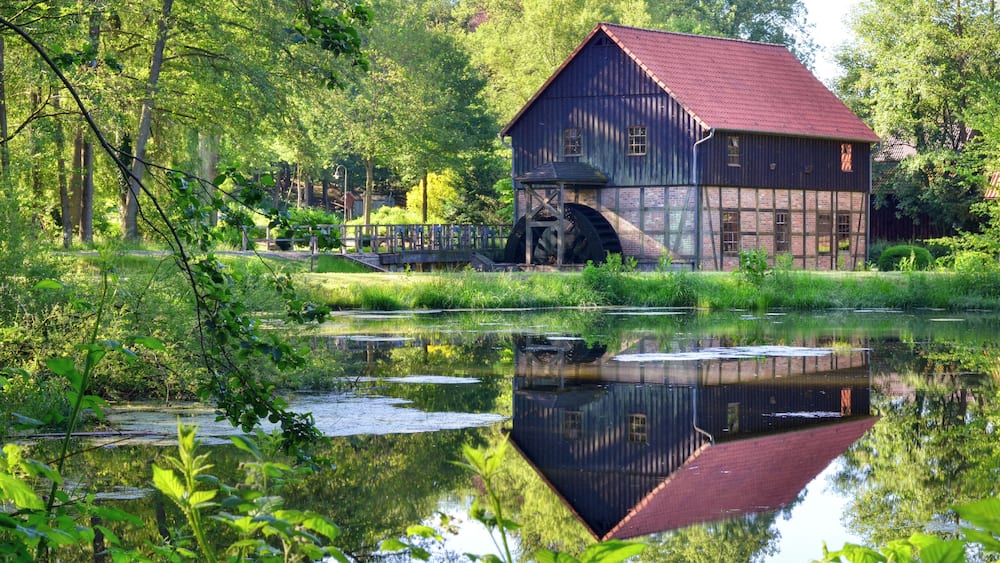 Eine hölzerne Mühle mit rotem Dach an einem ruhigen Fluss, umgeben von üppiger Vegetation.