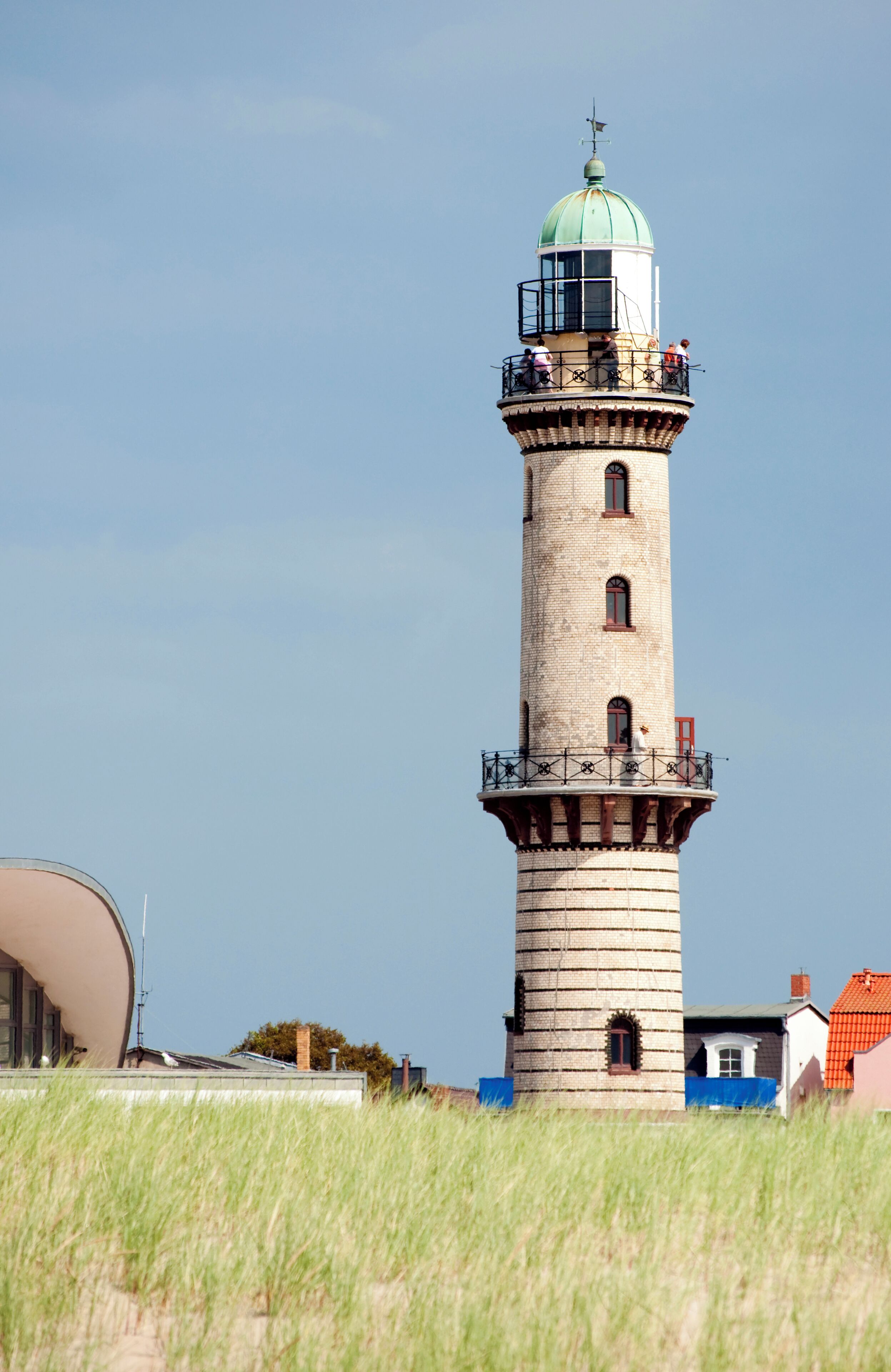 A tall lighthouse with a green dome and a white top, surrounded by grass and houses.
