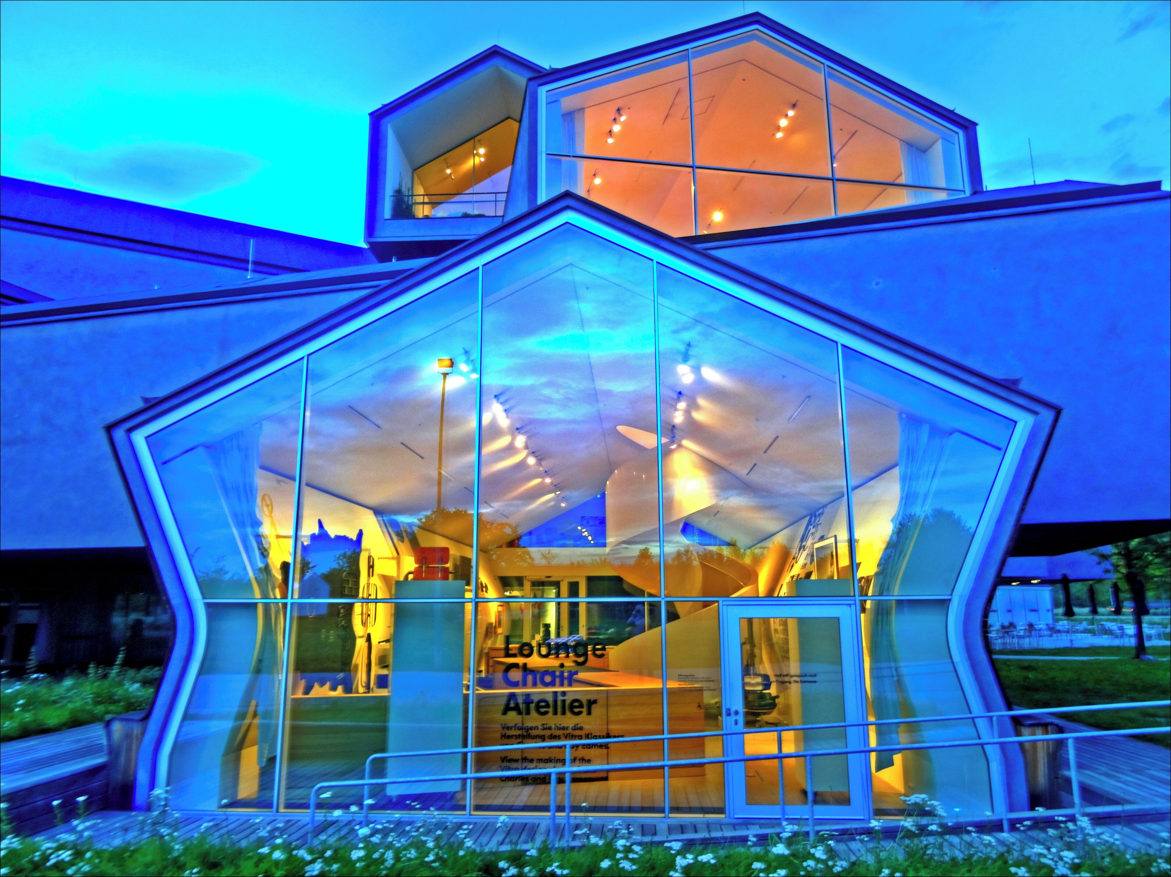 A modern glass building with a hexagonal structure and a sign that reads "Lounge Chair Atelier".