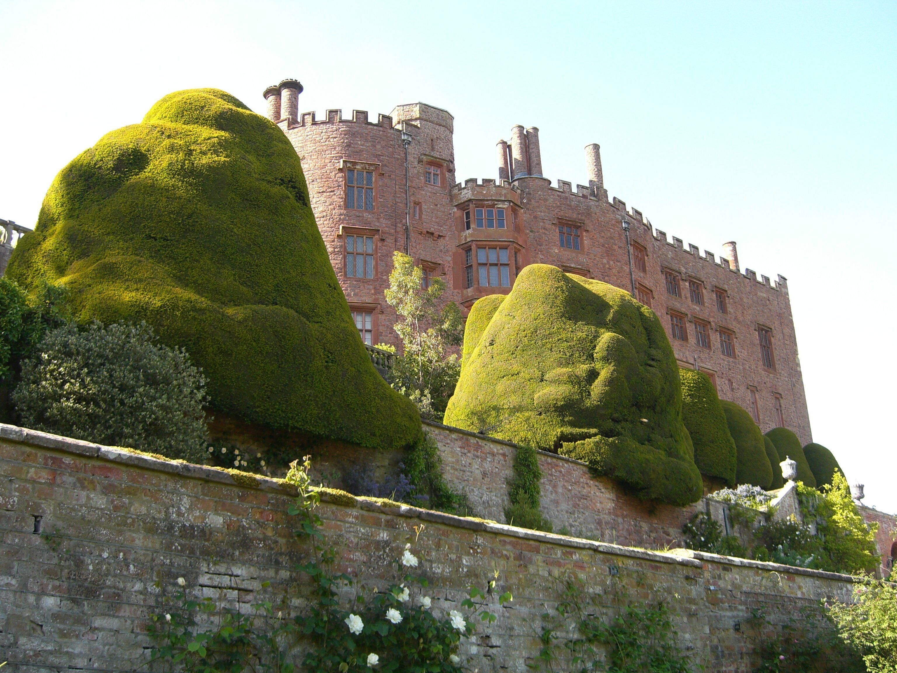 A castle with a stone facade and multiple chimneys. In front of the castle, there are large, topiary sculptures shaped like human figures.