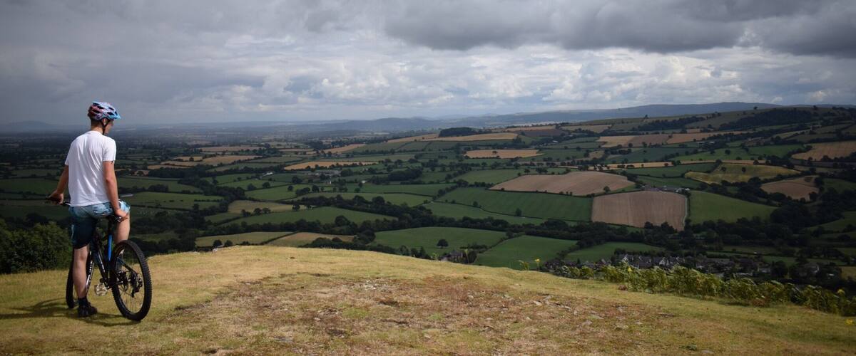 A cyclist on a hill overlooking a valley with fields and distant hills.