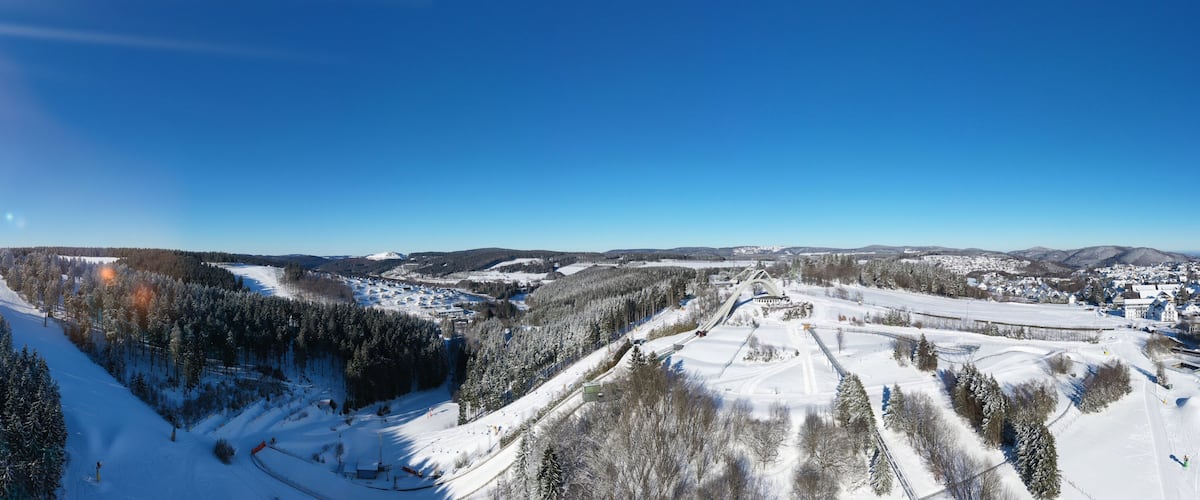 A snow-covered ski slope with a forested valley, a ski lift, and distant mountains.