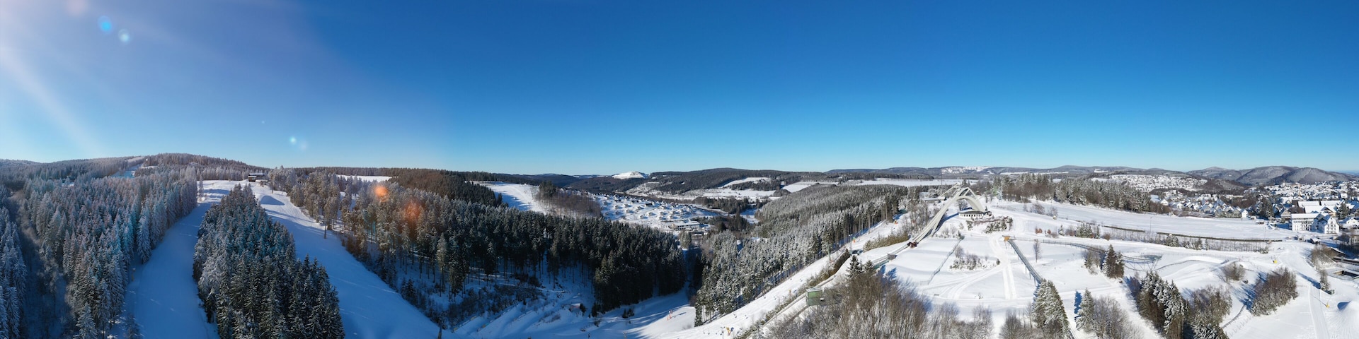 A snow-covered ski slope with a forested valley, a ski lift, and distant mountains.