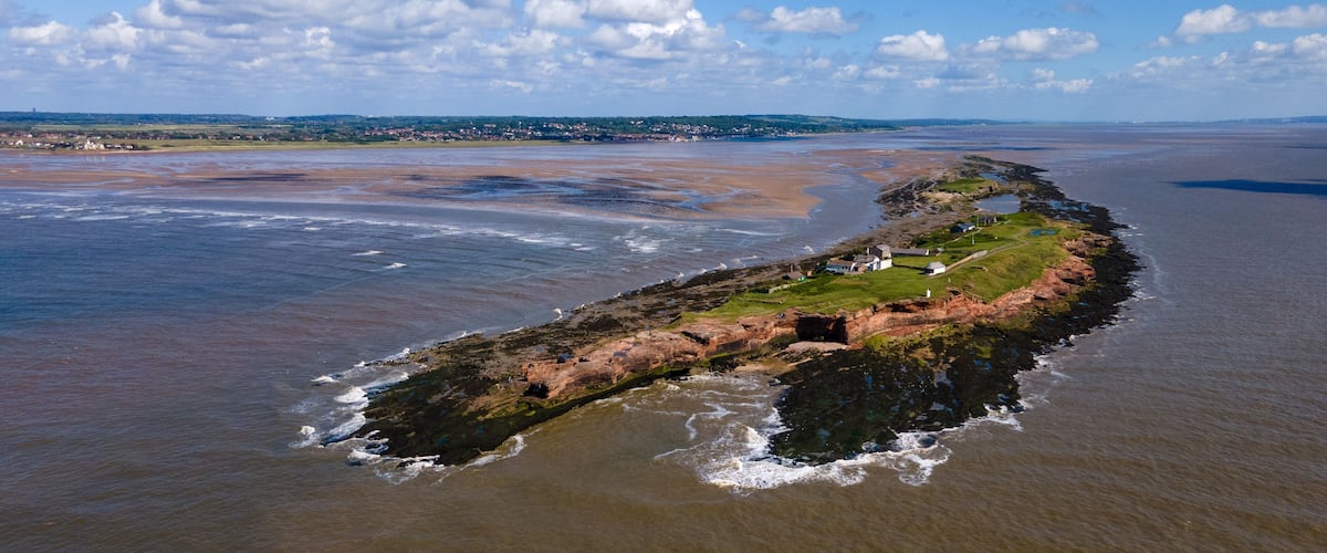 A coastal landscape with a rocky peninsula, a lighthouse, and a distant town.