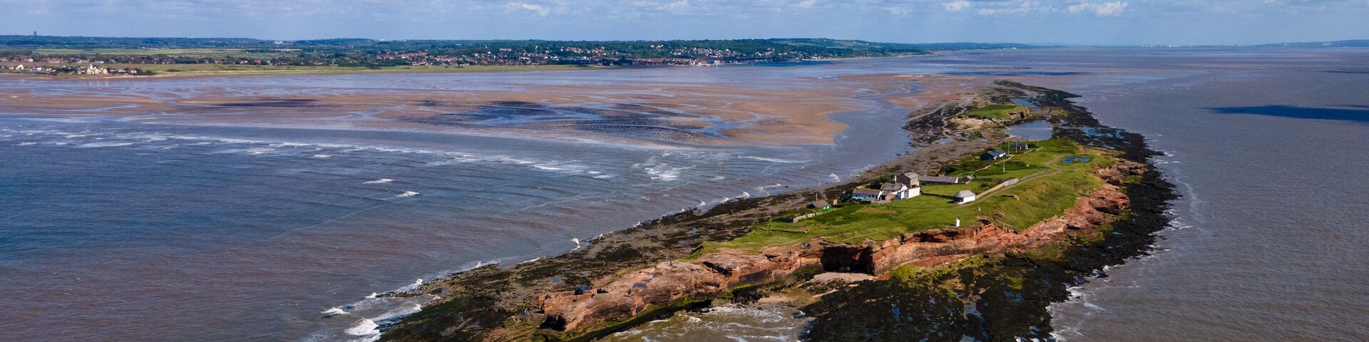 A coastal landscape with a rocky peninsula, a lighthouse, and a distant town.