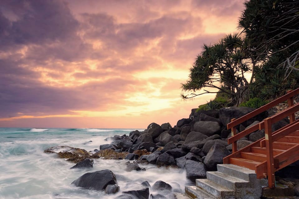 A rocky coastline with a wooden staircase leading down to the water, surrounded by trees and a sunset sky.