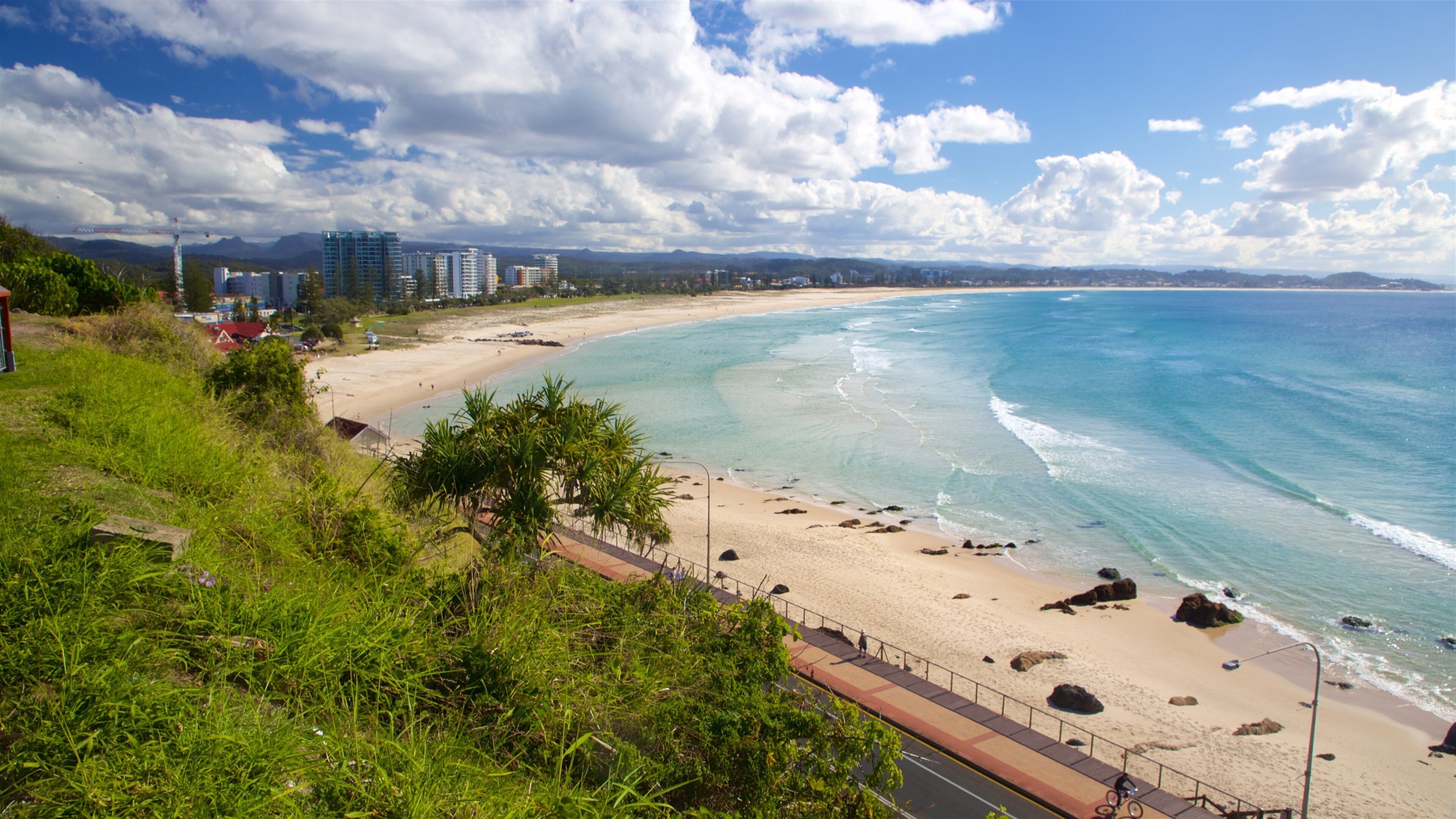 A coastal beach with clear turquoise waters, a sandy shore, and a backdrop of mountains and buildings.