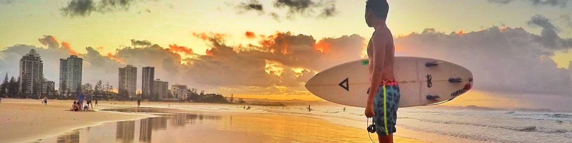 A beach with a sunset, a person with a surfboard, and buildings in the background.