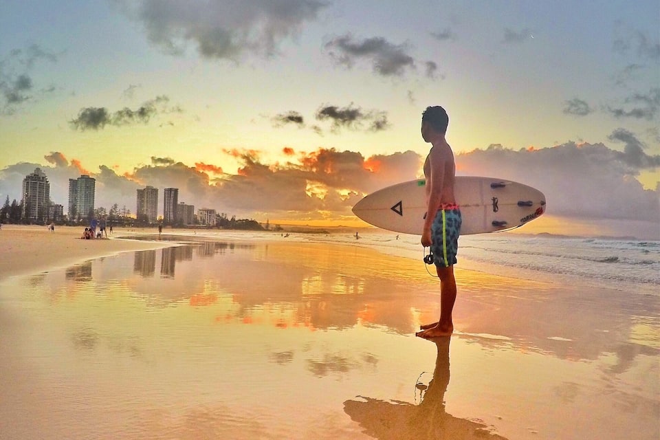 A beach with a sunset, a person with a surfboard, and buildings in the background.