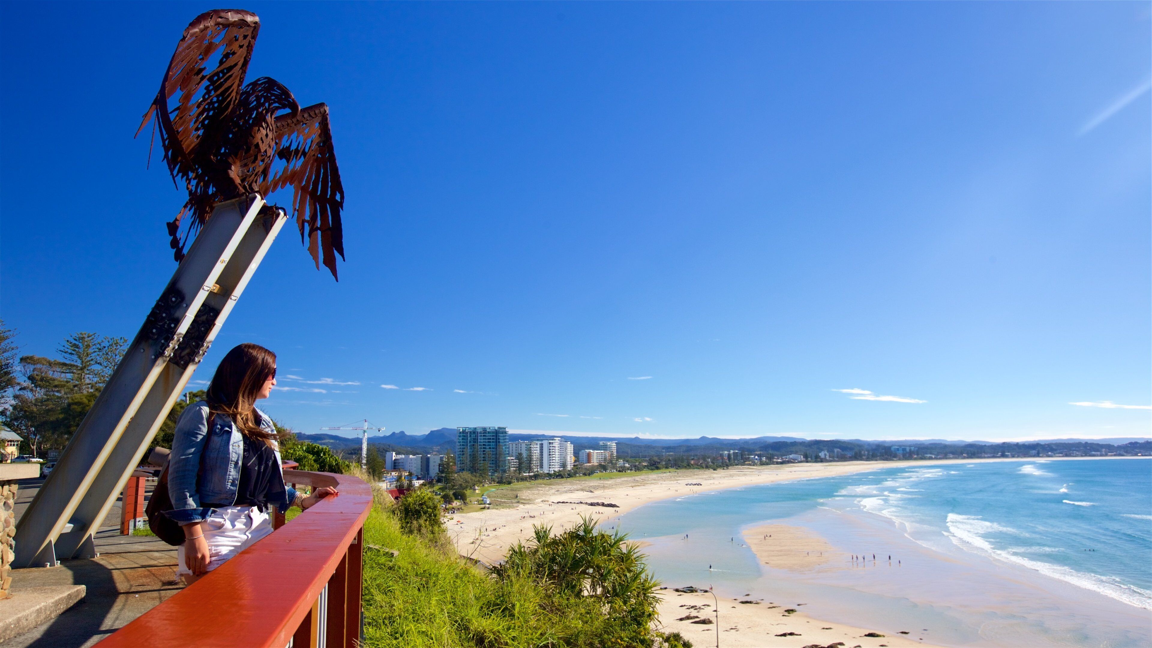 A beach with a metal sculpture, a person, and a cityscape in the background.