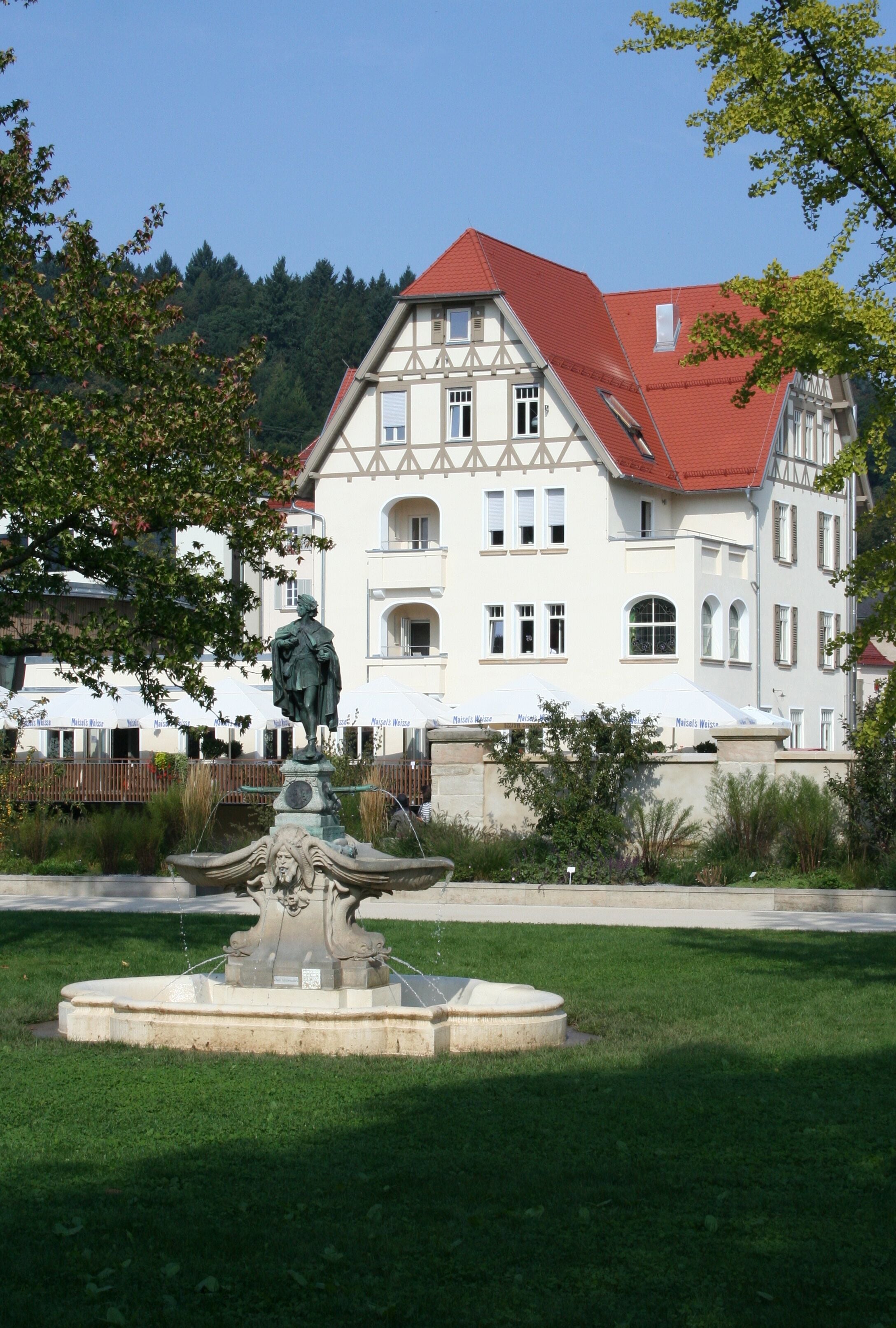 A white building with a red roof, a fountain, and trees in the foreground.