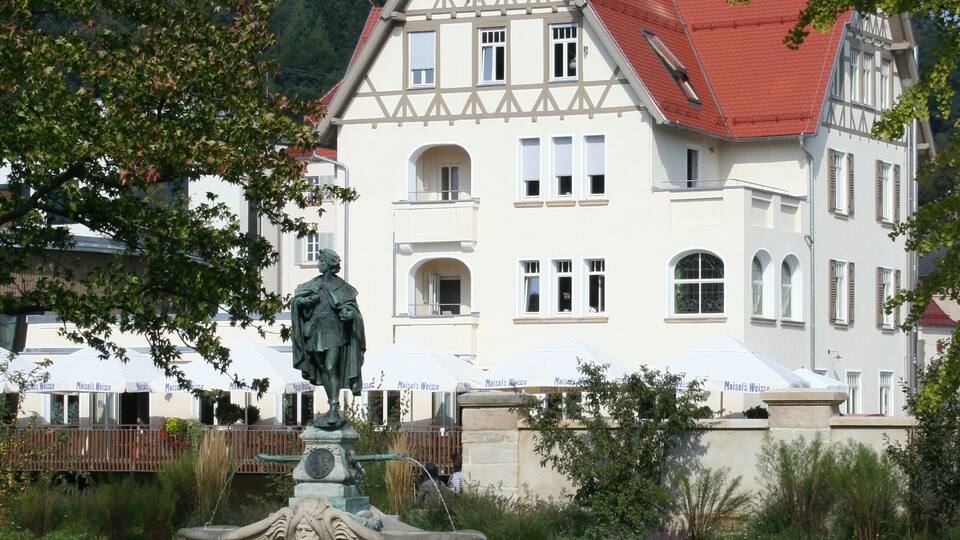A white building with a red roof, a fountain, and trees in the foreground.