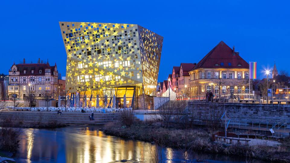 A cityscape at dusk with a modern building featuring star patterns, traditional architecture, and a river in the foreground.